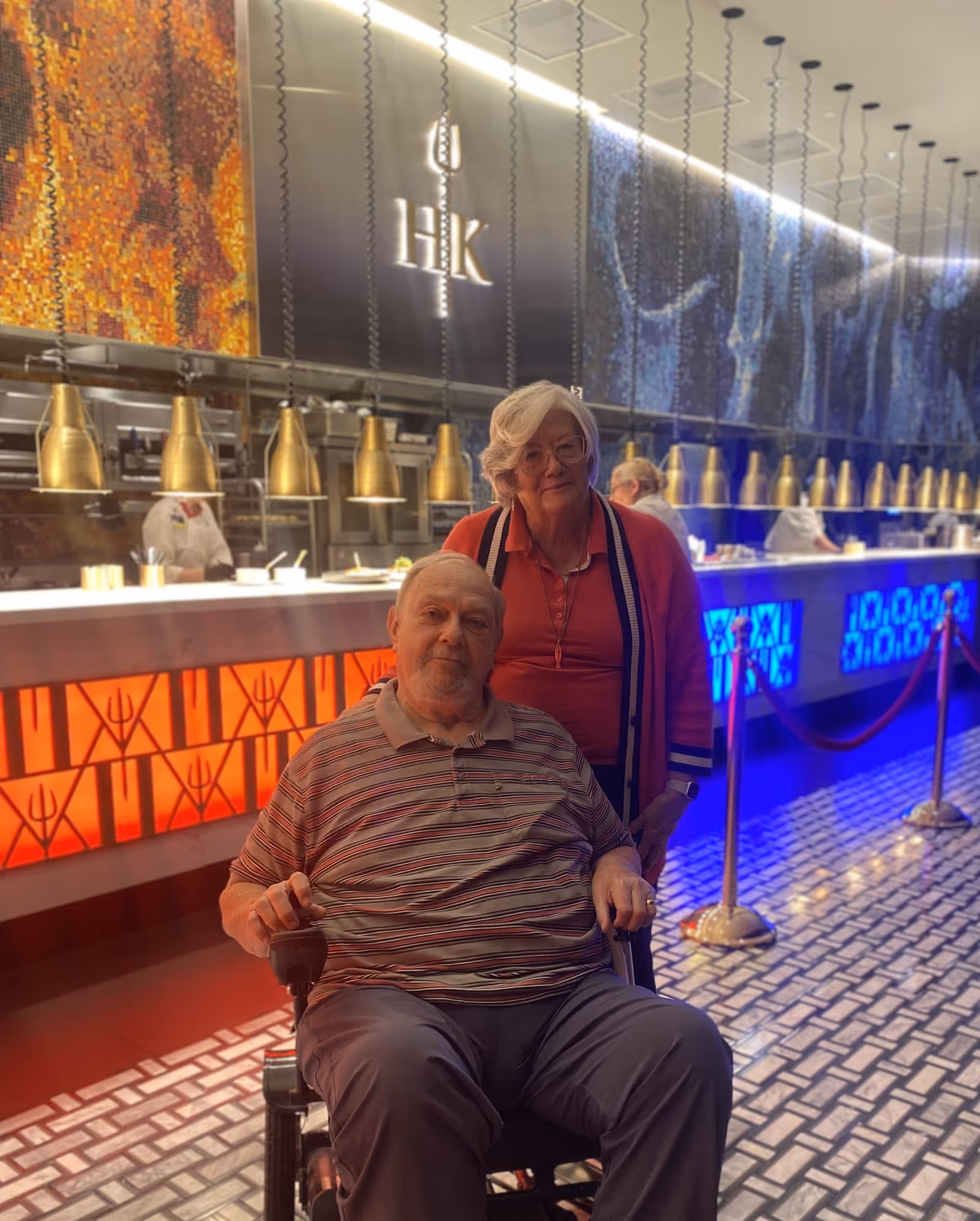 An elderly man in a wheelchair and a woman stand in front of a brightly lit open kitchen/food counter with blue and orange accent lighting.