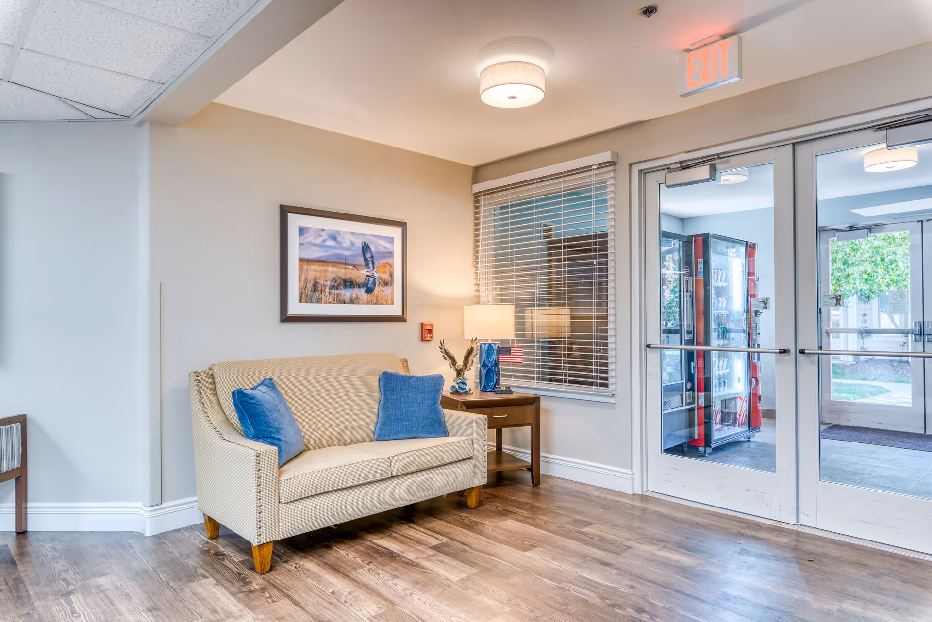 A cozy seating area in a senior living facility with a beige loveseat adorned with two blue pillows, a wooden side table with a lamp and decorative items, a framed picture of a bird in flight on the wall, and glass double doors leading outside where a vending machine is visible.