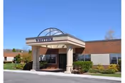 Exterior view of a single-story brick building with a covered entrance supported by columns. The entrance has a sign that reads 'WHITTIER'. There are shrubs and small trees planted around the building, and the sky is clear and blue.
