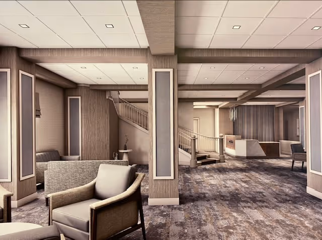 A spacious and modern interior common area with comfortable armchairs, carpeted floor, and a staircase leading to an upper level. The room features neutral tones with beige walls, recessed lighting in the ceiling, and a reception desk in the background.
