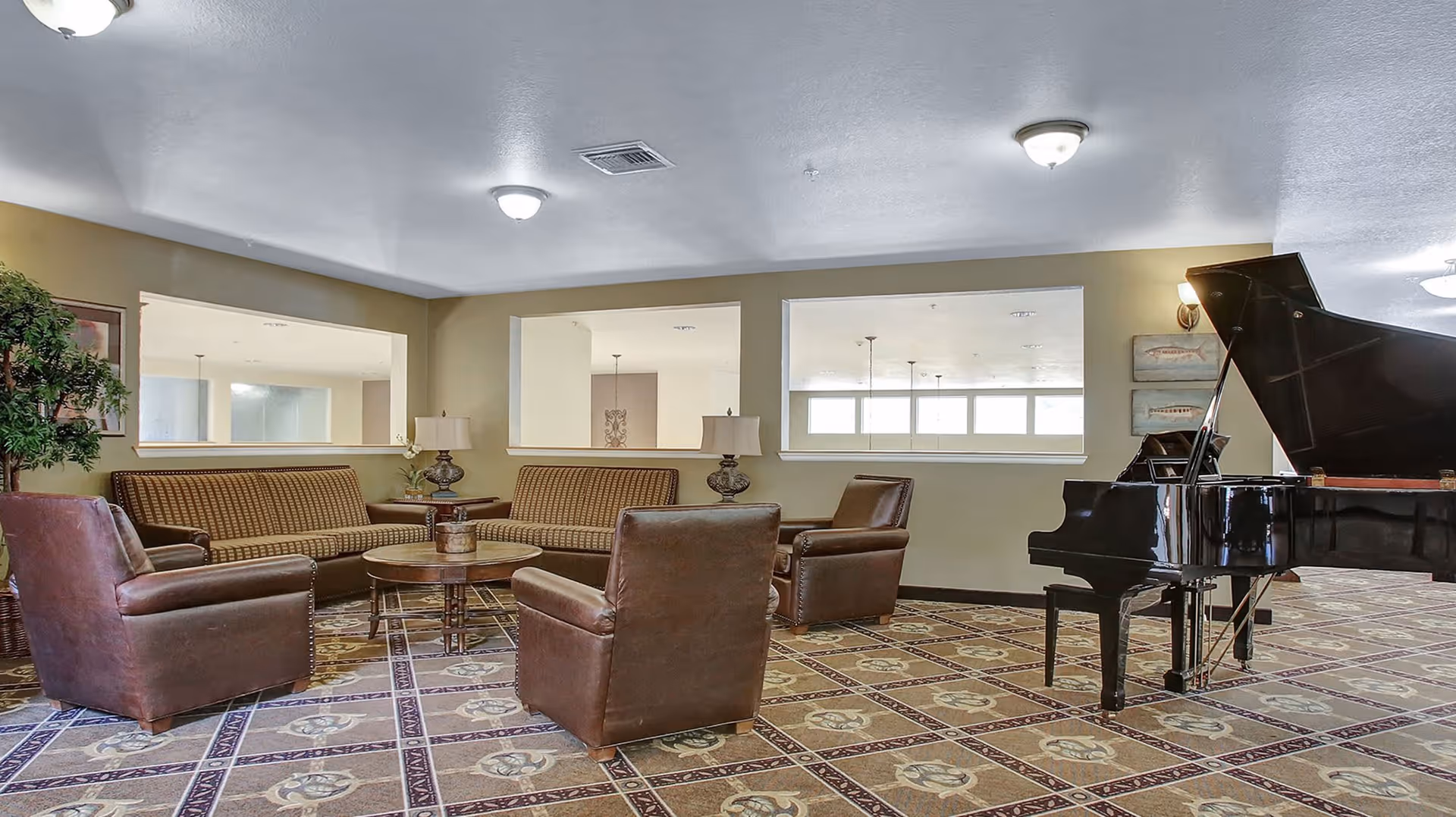 A senior living facility common area with two brown leather armchairs, two brown patterned sofas, a round wooden coffee table, and a black grand piano. The room has beige walls, patterned carpet flooring, and three large rectangular windows looking into another room.