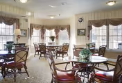 Bright dining room with round tables and chairs, large windows with draped valances, and potted plants.