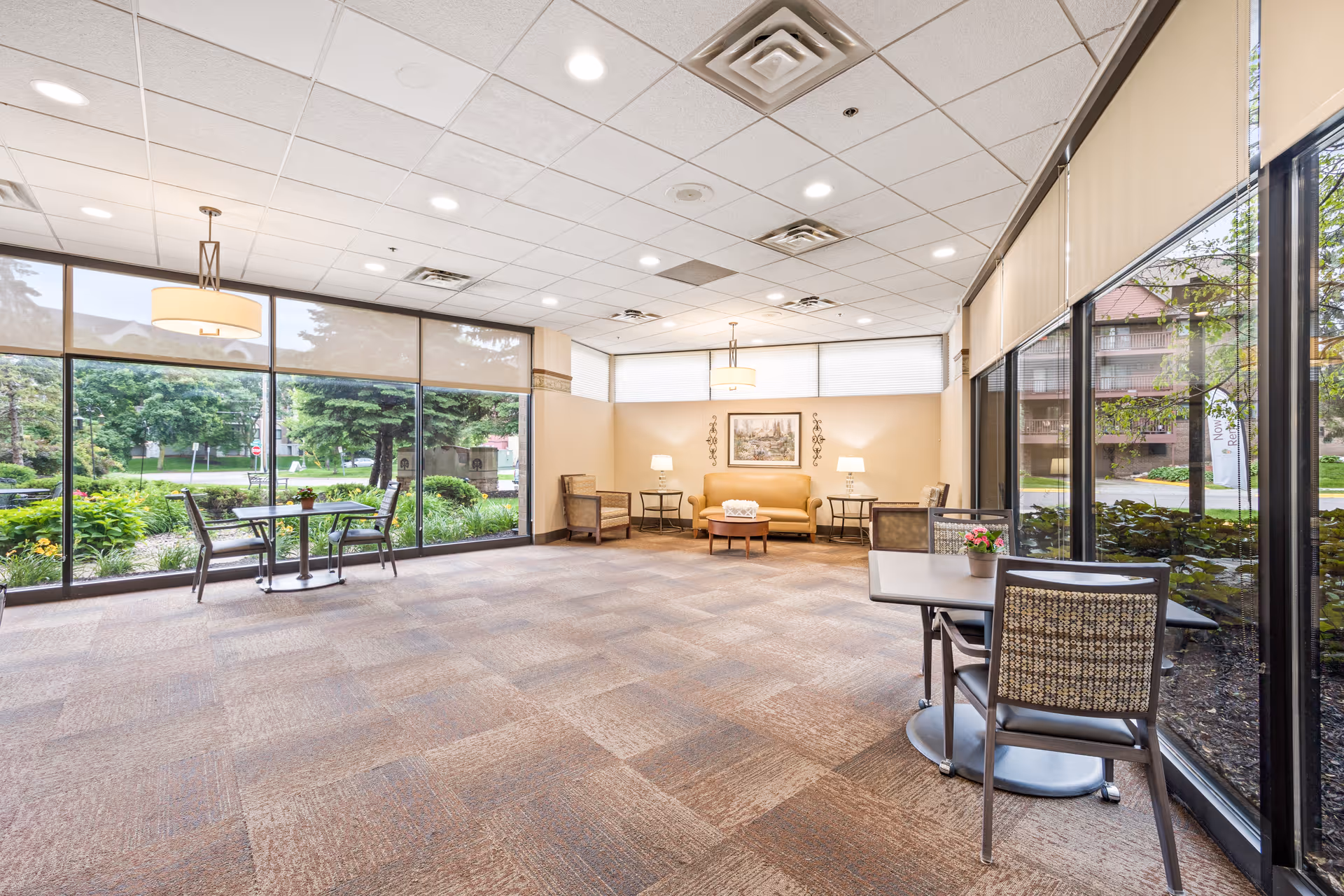 A bright and spacious senior living common area with large floor-to-ceiling windows showing greenery outside. The room features a beige couch, two armchairs, two small tables with lamps, and two small dining tables with chairs near the windows. The ceiling has recessed lighting and hanging light fixtures.