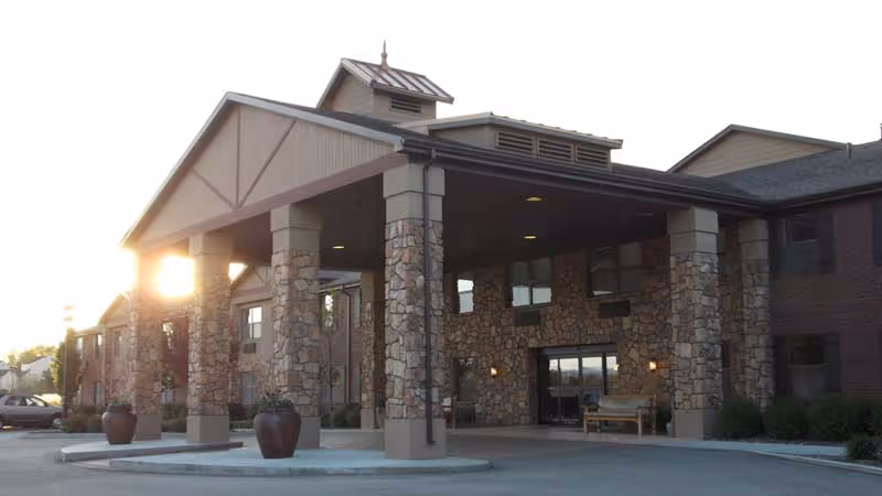 Exterior view of a senior living facility entrance with stone pillars supporting a covered driveway. The building has a combination of stone and brick facade with large windows and a bench near the entrance. The sun is setting in the background.