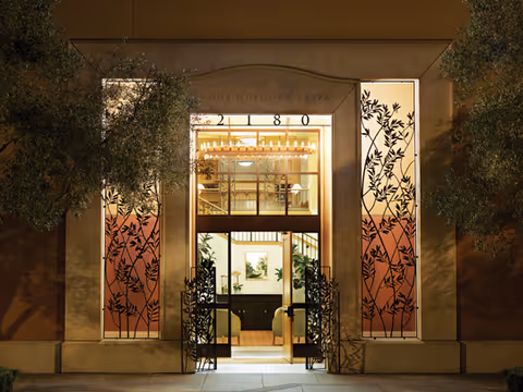 Night view of the entrance to Rhoda Goldman Plaza, a building with illuminated glass panels featuring decorative leaf patterns on either side of the doorway, flanked by trees.