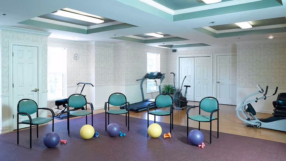 A fitness room with exercise equipment including a treadmill, stationary bike, and weight bench. Four green cushioned chairs are arranged in a semi-circle on a purple mat, each accompanied by small exercise weights and stability balls. The room has light-colored walls, windows, and a ceiling with recessed lighting.