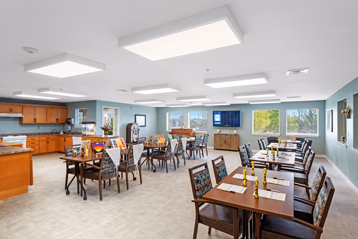 A spacious dining room in a senior living facility with multiple wooden tables and chairs arranged neatly. Each table has menus and yellow condiment bottles. The room has large windows letting in natural light, a wall-mounted TV, and a kitchen area with wooden cabinets and appliances in the background. The walls are painted light blue, and the ceiling has several rectangular light fixtures.