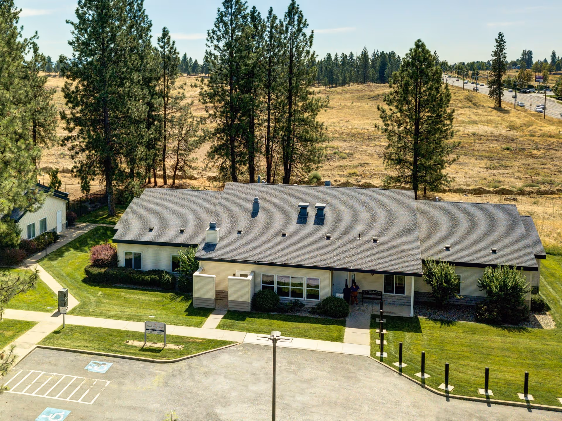 Aerial view of a single-story building with a gray roof surrounded by green lawns and trees. The building is part of North Point Village facility, with a parking lot in the foreground and a road visible in the background. The area around the building includes paved walkways and landscaped bushes.