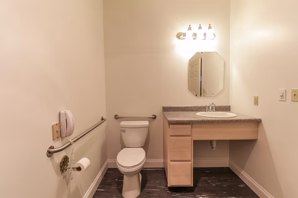 A clean and simple bathroom with a white toilet, a wall-mounted grab bar, a mounted telephone, a toilet paper holder, a vanity with a sink and countertop, an octagonal mirror, and a light fixture above the mirror. The walls are painted white and the floor has dark wood-like tiles.