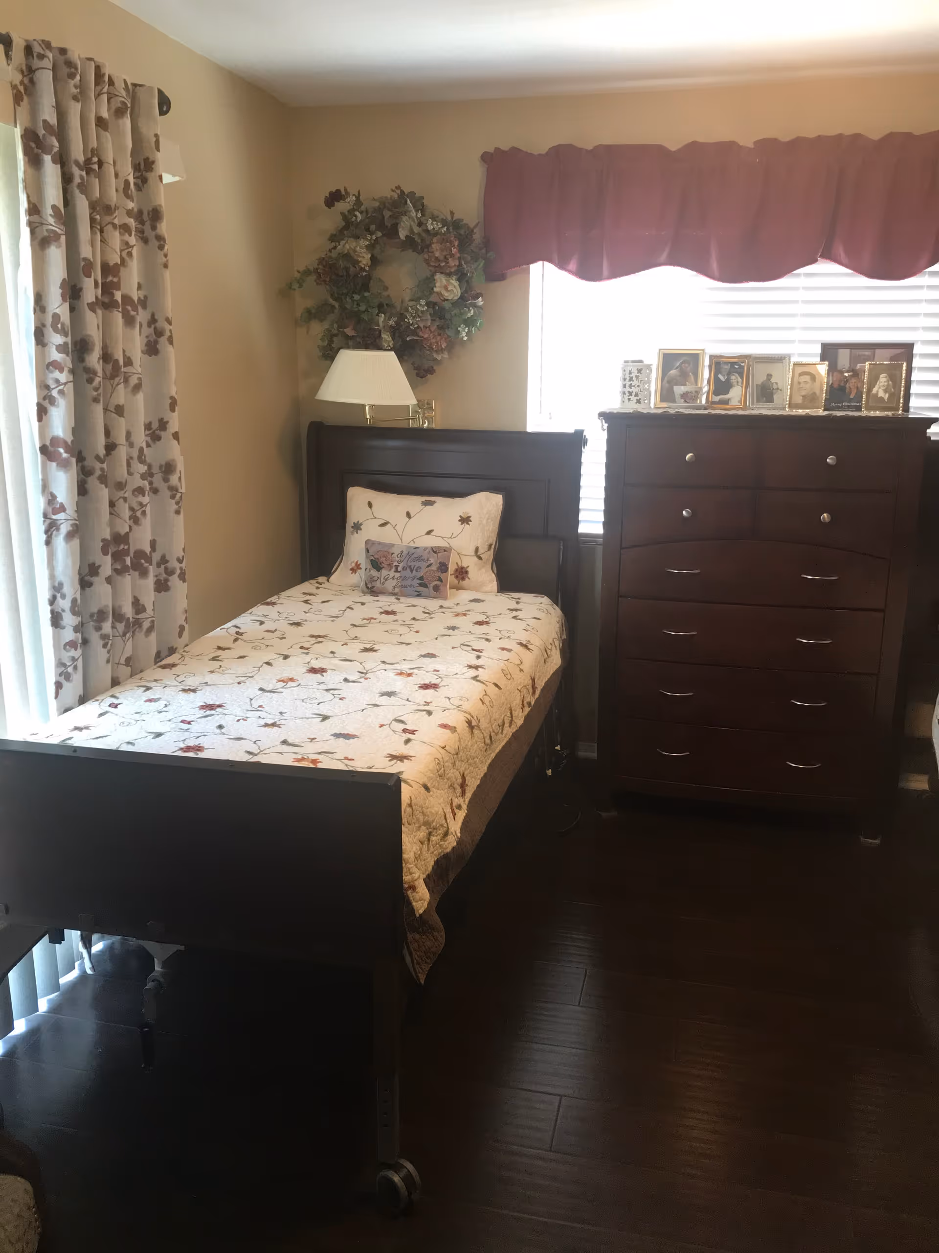 A cozy bedroom with a single bed covered in a floral quilt and matching pillow. There is a wooden dresser with several framed photos on top, a lamp on a small table next to the bed, floral curtains on the window, and a decorative wreath hanging on the wall.