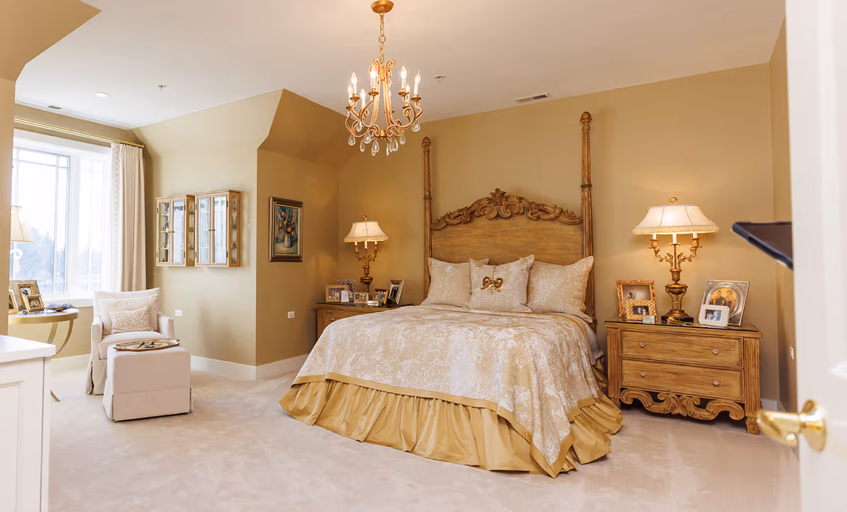 Elegant traditionally decorated bedroom with an ornate wooden bed, matching nightstands and lamps, a chandelier, and a window-side armchair.