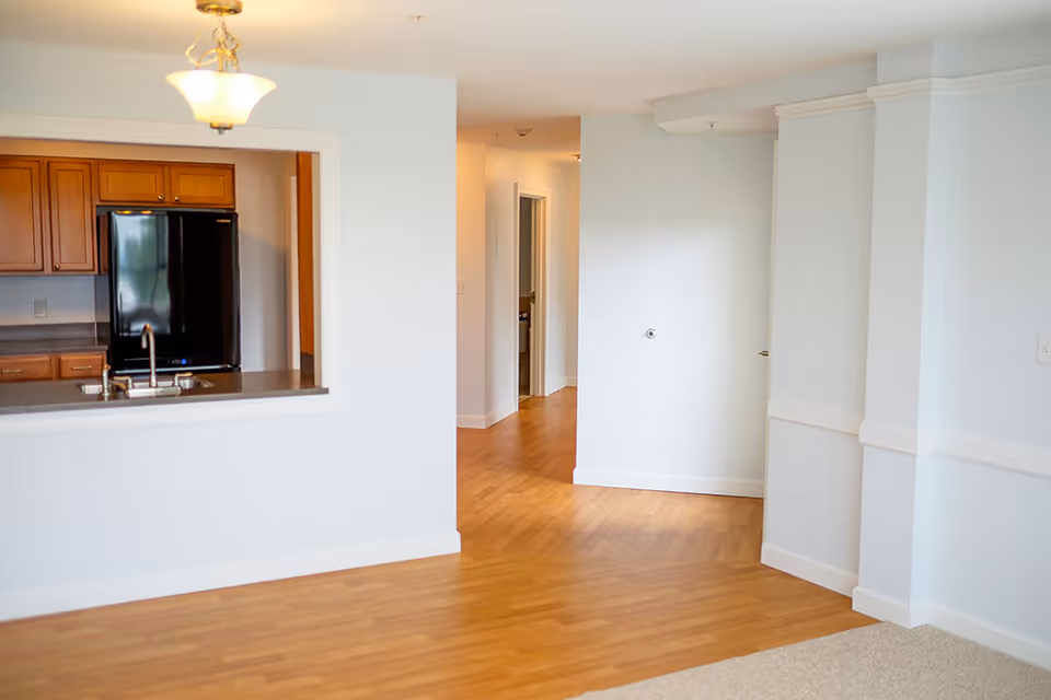 Interior view of a senior living facility showing a kitchen area with wooden cabinets and a black refrigerator visible through a pass-through window. The room has light-colored walls, wooden flooring, a ceiling light fixture, and a hallway leading to other rooms.