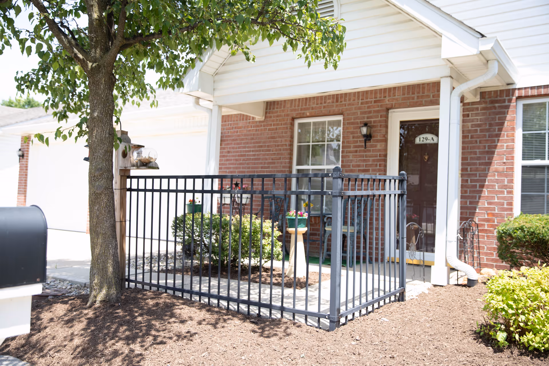 Front porch area of a brick residential unit with a black metal railing enclosing a small patio. There is a tree and a mailbox in the foreground, and the door has a sign with the number 129-A. Some potted plants and bushes are visible around the porch.