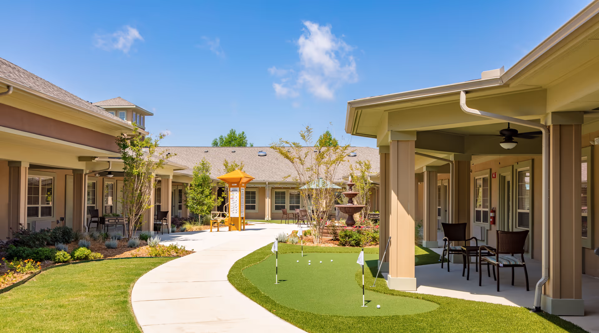 Outdoor courtyard area at Village on the Park McKinney featuring a putting green with golf flags and balls, a paved walkway, patio seating with chairs and tables under covered areas, landscaped plants and trees, and a water fountain in the background under a clear blue sky.