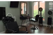 Salon styling area with chairs, a hair-washing sink, mirrors, a standing hair dryer, and windows overlooking brick buildings.