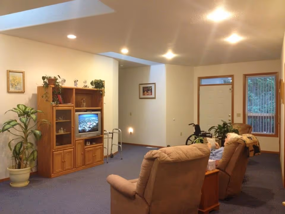 A cozy living room with beige recliner chairs, a wooden entertainment center with a TV, potted plants, framed pictures on the walls, and a walker near the entertainment center. A person is sitting in one of the recliners reading a newspaper. The room has blue carpet and recessed ceiling lights.