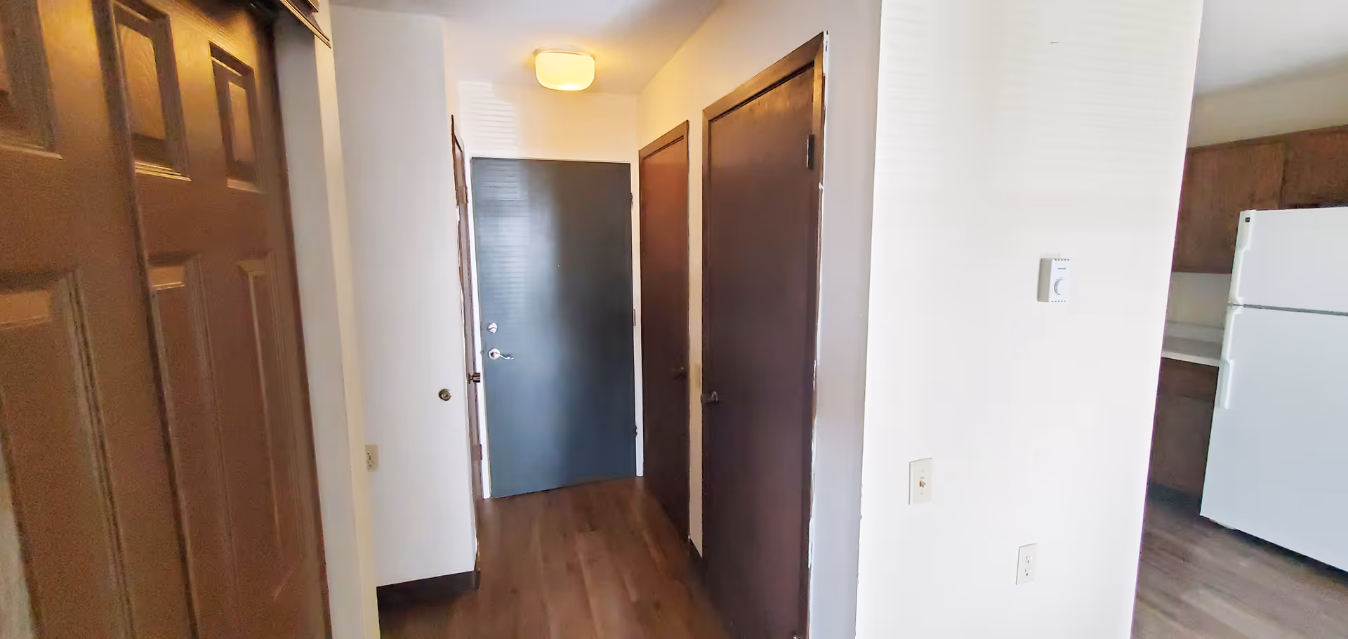 A hallway in an apartment with a dark blue front door at the end, two brown doors on the right side, and a brown door on the left side. The floor is wooden, and part of a kitchen with a white refrigerator and brown cabinets is visible on the right.