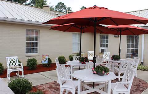 Outdoor patio area with white chairs and tables arranged under large red umbrellas. The patio is surrounded by a beige brick building with windows and some small green bushes and plants along the building's edge.