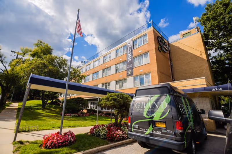 Exterior view of a multi-story brick building with a covered entrance and a flagpole with an American flag. There are well-maintained flower beds and greenery around the entrance. A black van with green and white lettering for The Grand Rehabilitation And Nursing At Queens is parked near the entrance under the canopy. The sky is partly cloudy with blue patches.