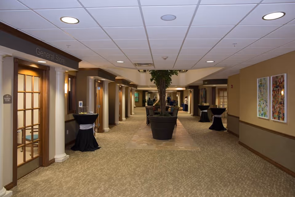 Interior hallway of Cahaba Ridge Retirement Community with carpeted floor, beige walls, and ceiling lights. There are several doors along the hallway, including one labeled 'General Store'. In the center, there is a large potted plant with chairs around it. Small round tables covered with black cloth and white sashes are placed along the hallway. Two framed artworks hang on the right wall.
