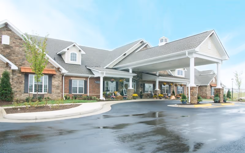 Exterior view of a senior living facility building with a covered entrance, stone and brick facade, windows with shutters, and a wet driveway in front.