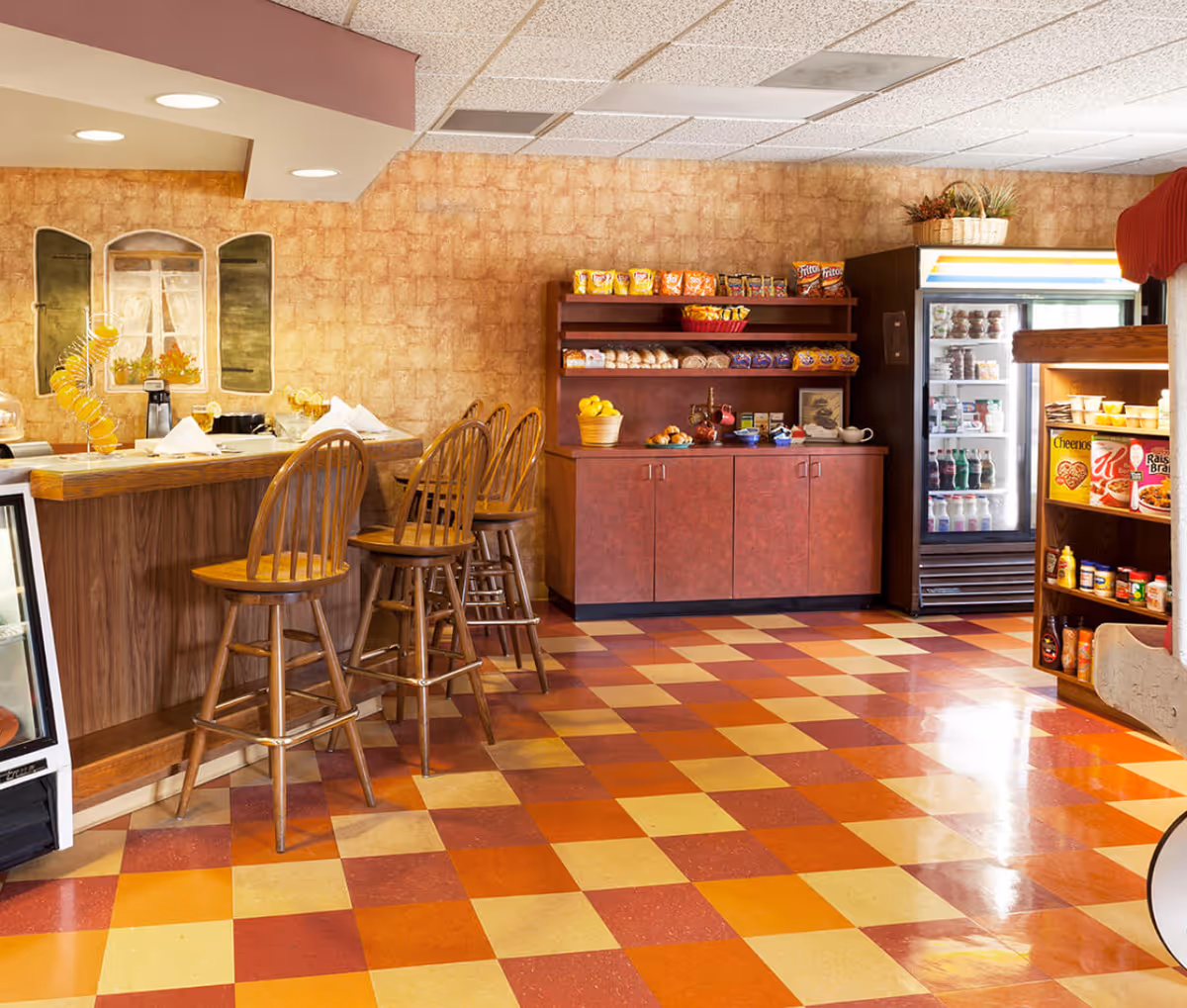 Small snack bar and seating area with wooden bar stools, shelves of packaged snacks, and a refrigerated display in a warm-toned interior.