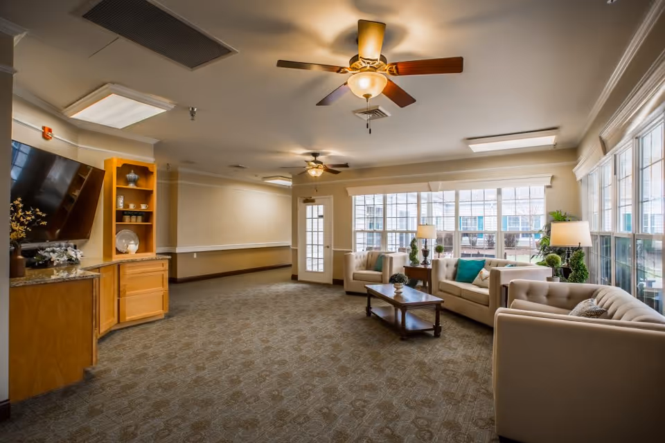 A spacious and well-lit living room area in Charter Senior Living of Franklin featuring beige sofas with colorful cushions, a wooden coffee table, large windows allowing natural light, ceiling fans with lights, and a wooden cabinet with a mounted flat-screen TV.