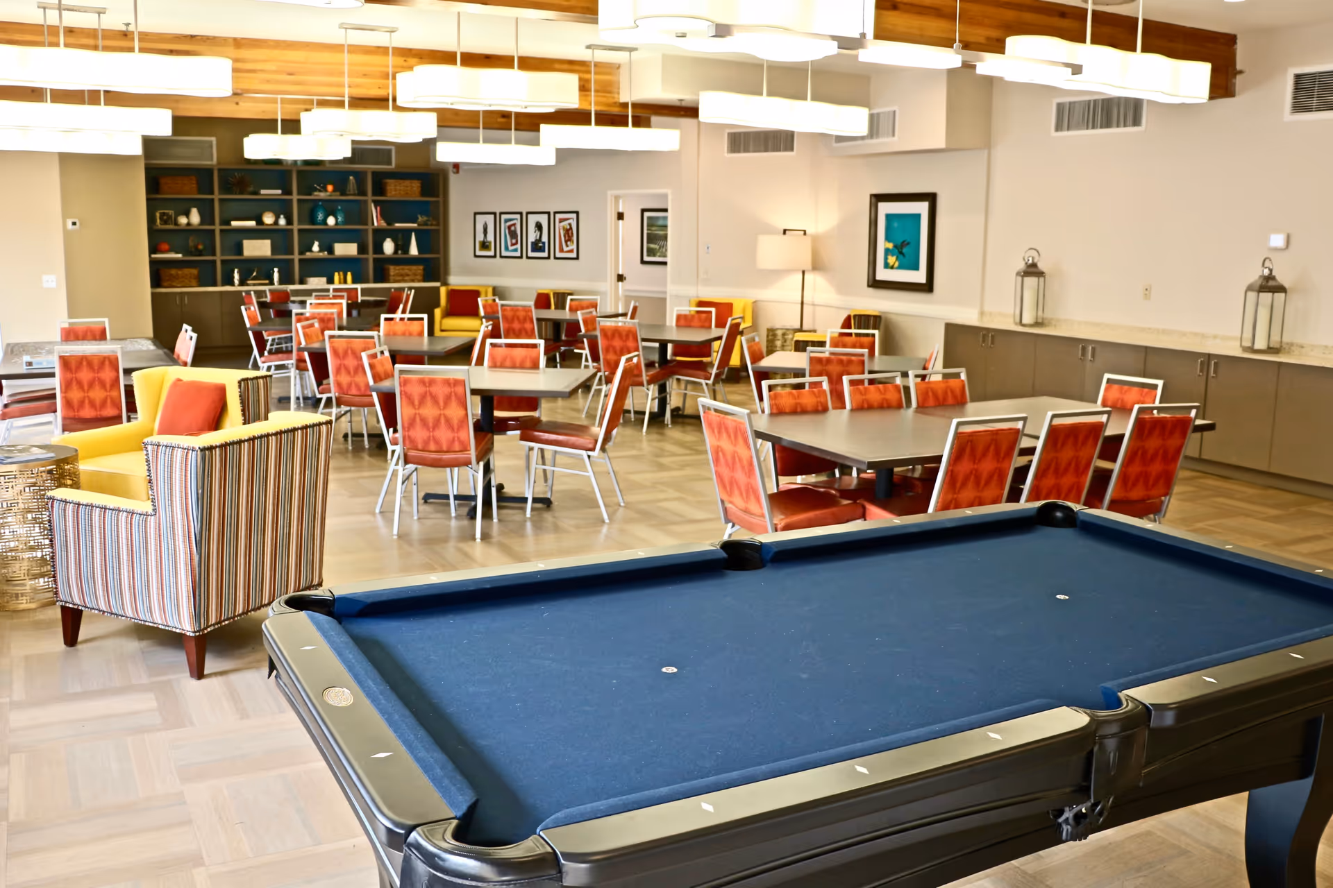 A spacious senior living common area featuring a blue pool table in the foreground, multiple tables with red cushioned chairs, striped and yellow armchairs, modern ceiling lights, and decorative shelves and artwork on the walls.