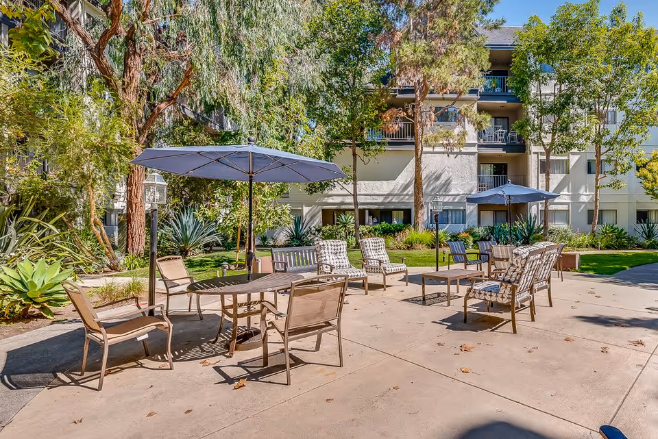 Outdoor patio area at Westminster Terrace Senior Living with multiple seating arrangements including chairs and tables under large umbrellas, surrounded by trees and greenery with a multi-story building in the background.