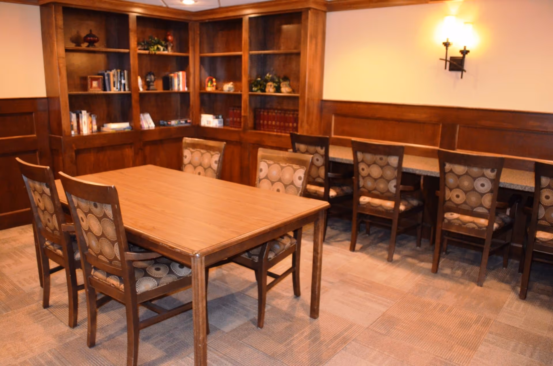 A room with a wooden table surrounded by six chairs with patterned upholstery. Behind the table, there is a long counter with several chairs lined up against the wall. The walls have wooden paneling and built-in bookshelves filled with books and decorative items. The room is softly lit with wall-mounted lights.