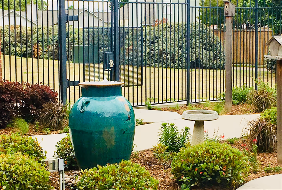 Outdoor garden area with a large green ceramic water fountain, a birdbath, various bushes and plants, a paved walkway, and a black metal fence in the background.
