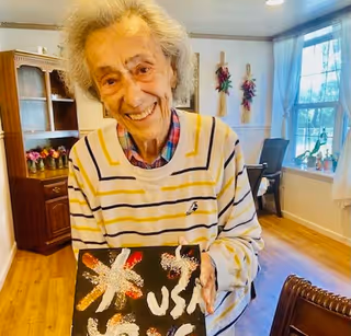 An elderly person smiling and holding a piece of artwork with colorful designs and the text 'USA' in a well-lit room with wooden flooring, a window with white curtains, and furniture including a wooden cabinet and chairs.