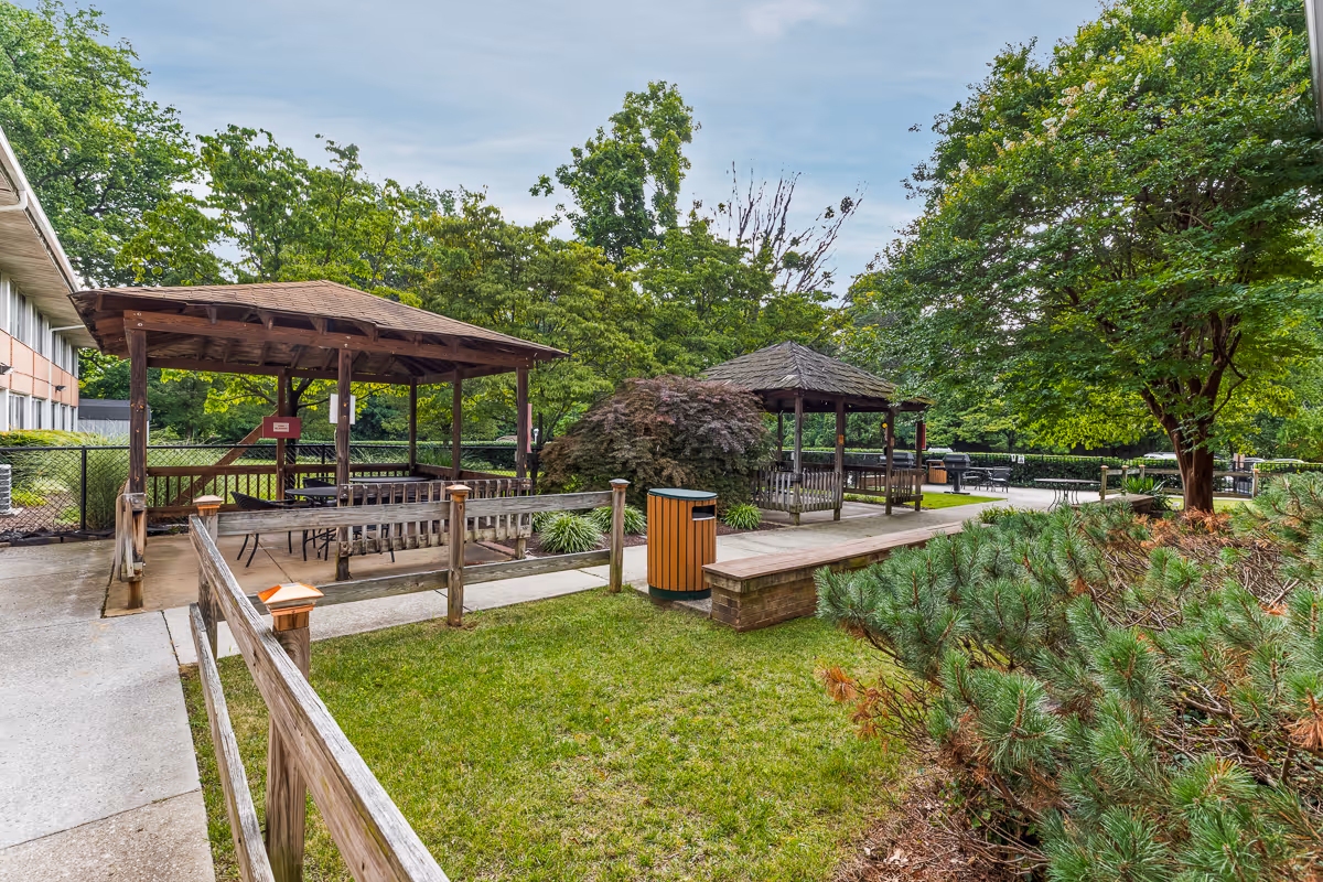 Outdoor area at Autumn Lake Healthcare at Arcola featuring two wooden gazebos with seating, surrounded by green grass, trees, and shrubs. There is a paved walkway, wooden railings, and a trash bin in the center. The sky is partly cloudy.