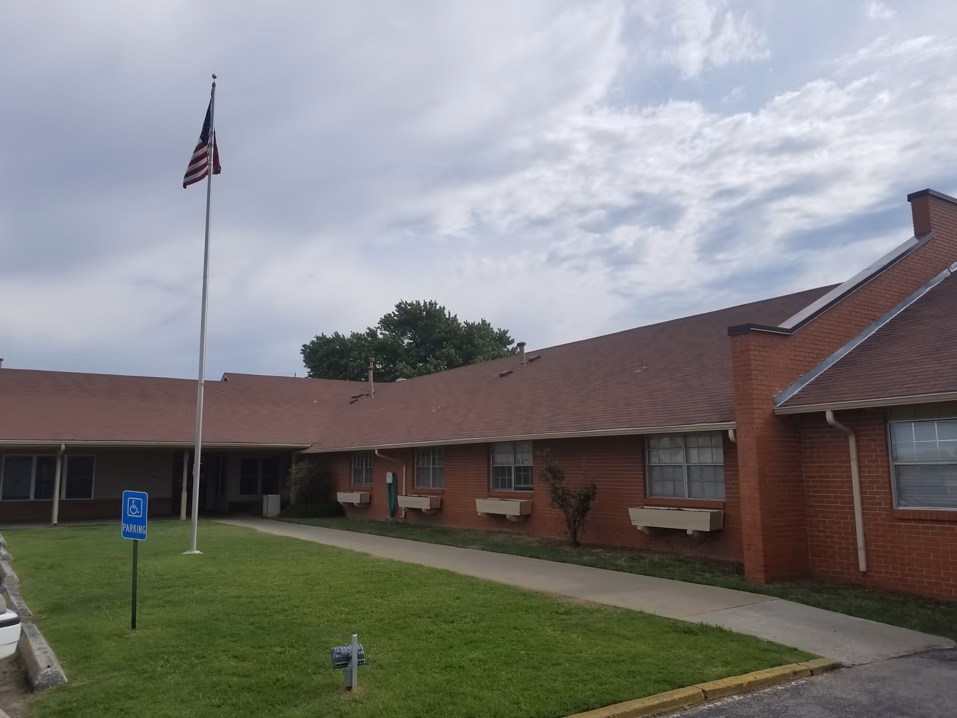 Exterior view of a single-story brick building with a sloped roof, an American flag on a tall flagpole, a sidewalk leading to the entrance, and a grassy area with a handicap parking sign.