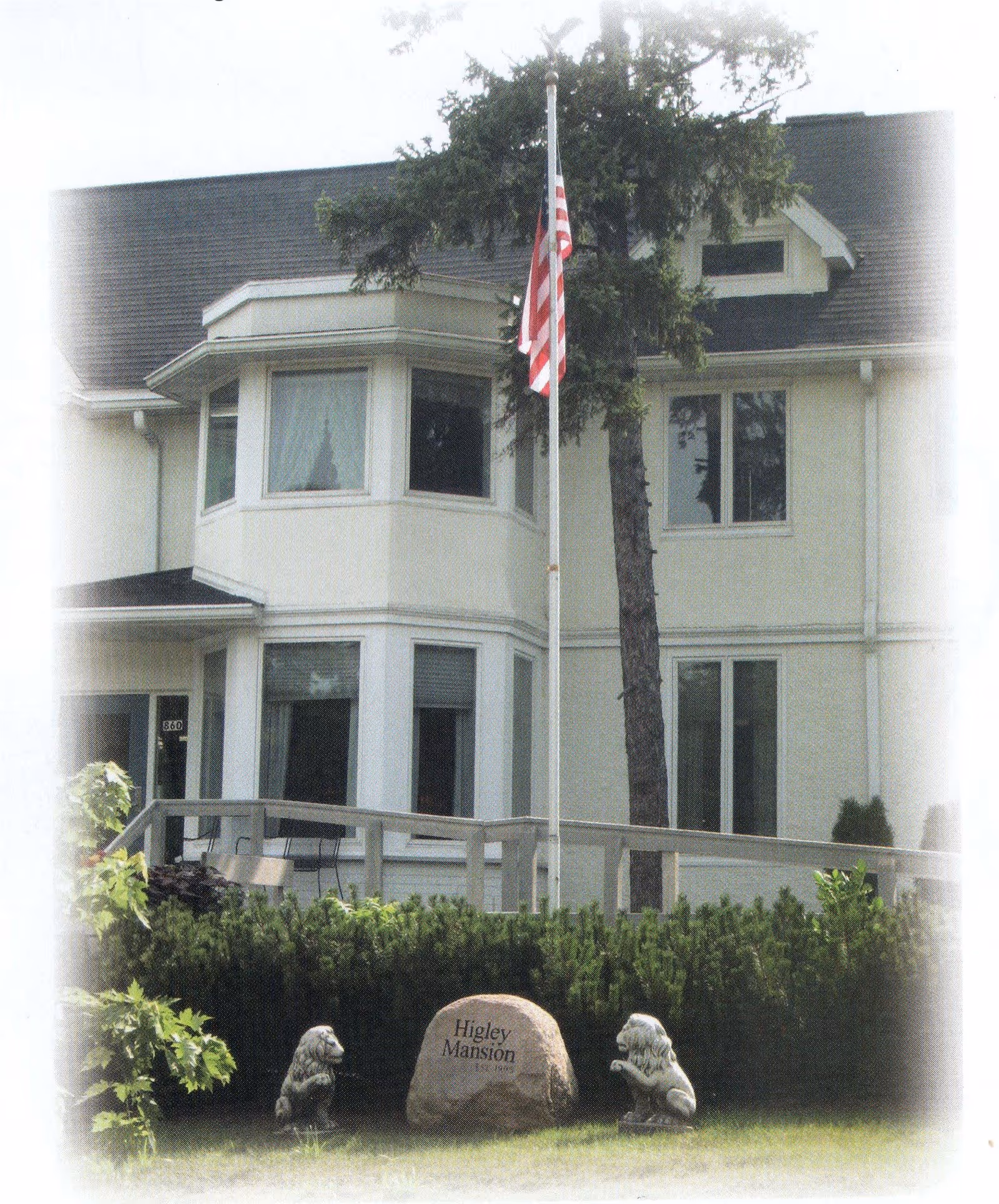 Two-story light-colored mansion front with an American flag, a stone engraved "Higley Mansion", and two small lion statues on the lawn.