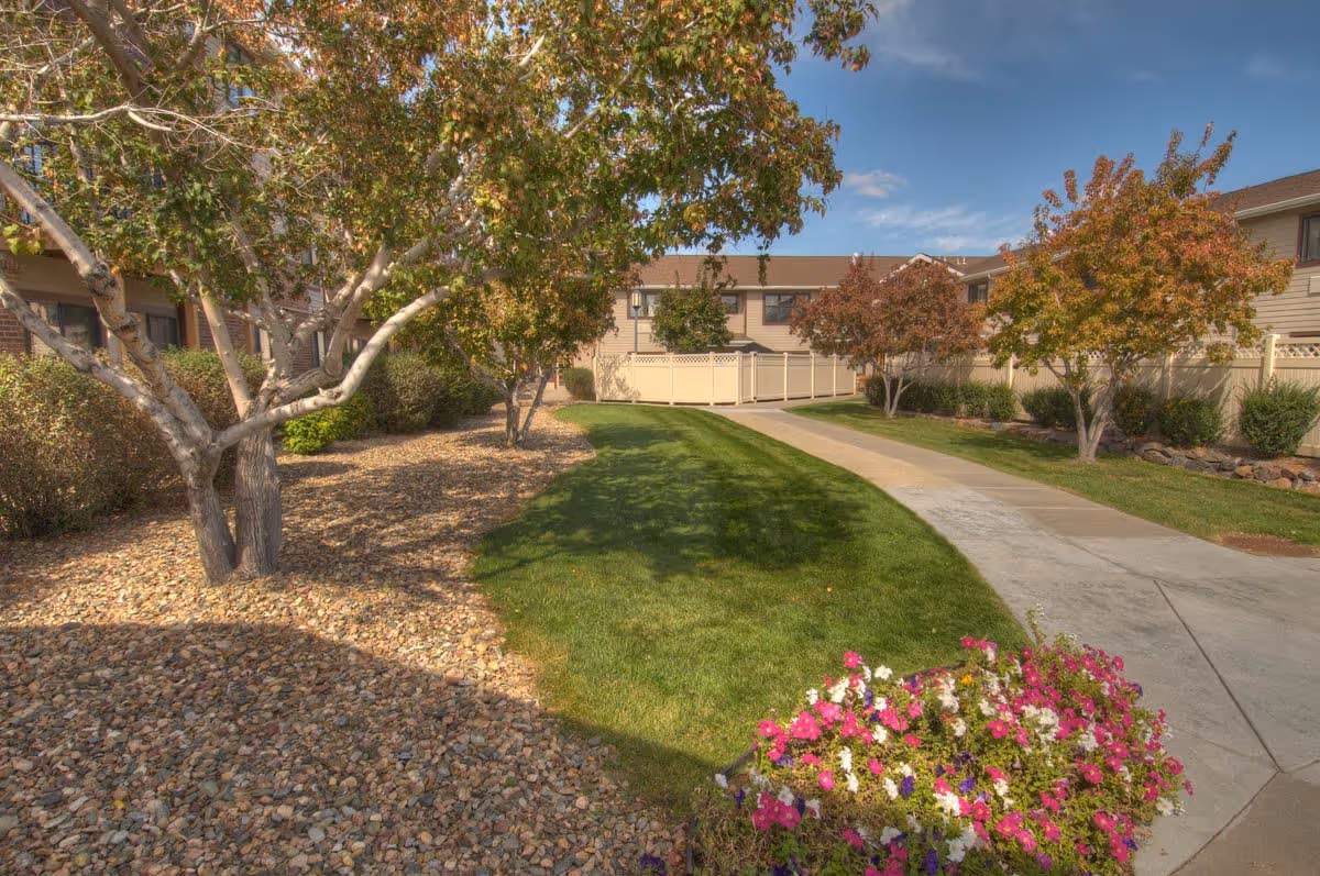Outdoor pathway in a senior living facility with green grass, trees with autumn foliage, and colorful flowers in the foreground. Residential buildings with beige siding and brown roofs are visible in the background under a partly cloudy sky.