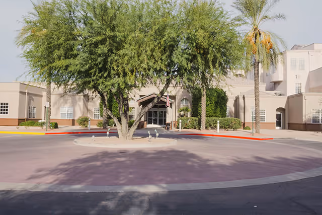 Front exterior view of Brookdale Baywood facility showing a circular driveway with a large tree and palm tree in the center, beige building with multiple windows, and an entrance with a covered awning.