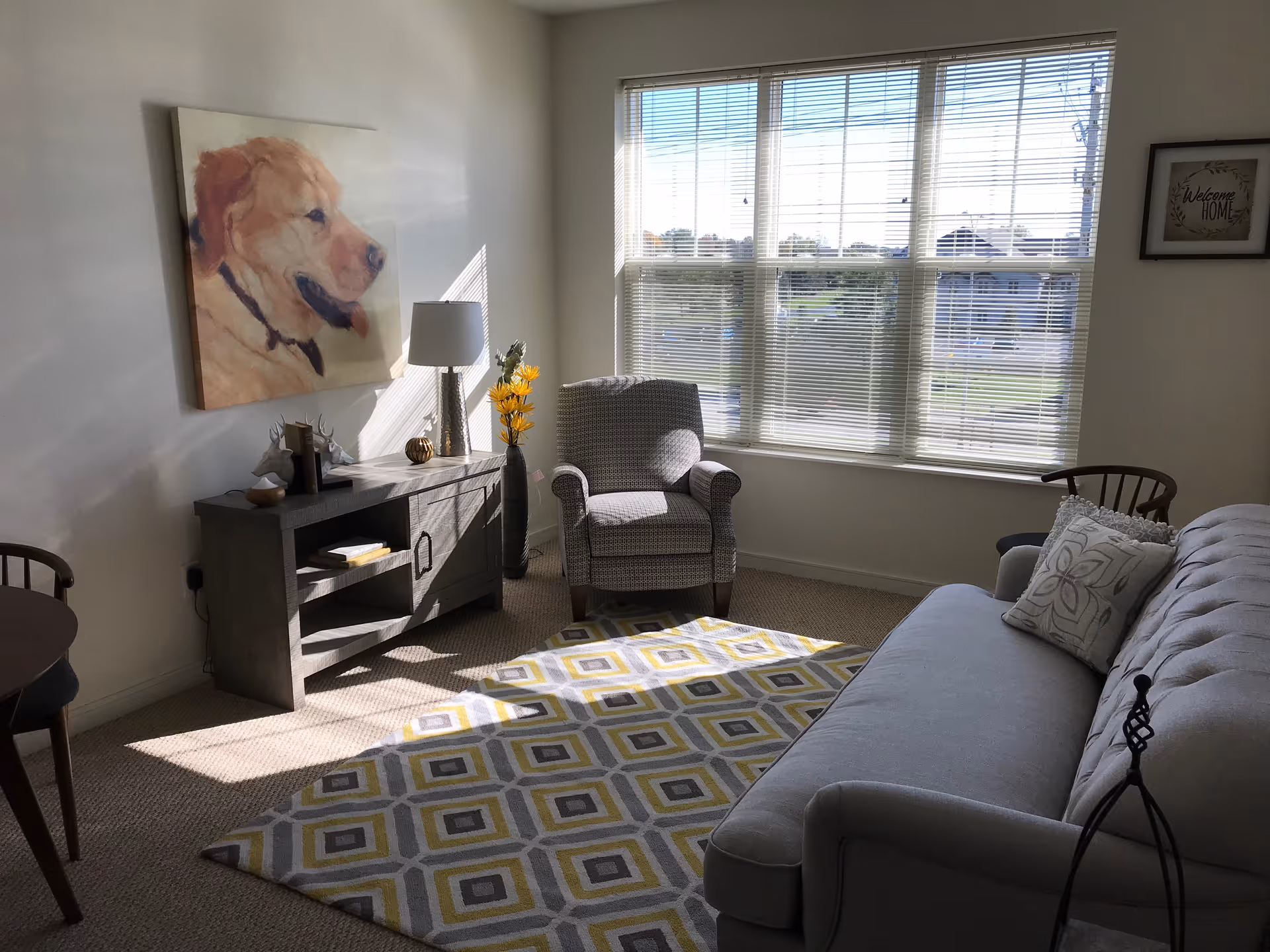 A bright living room with a large window letting in natural light. The room features a gray upholstered sofa with decorative pillows, a patterned armchair, a wooden console table with a lamp, books, and decorative items, and a colorful geometric area rug. A large painting of a golden retriever dog hangs on the wall above the console table.