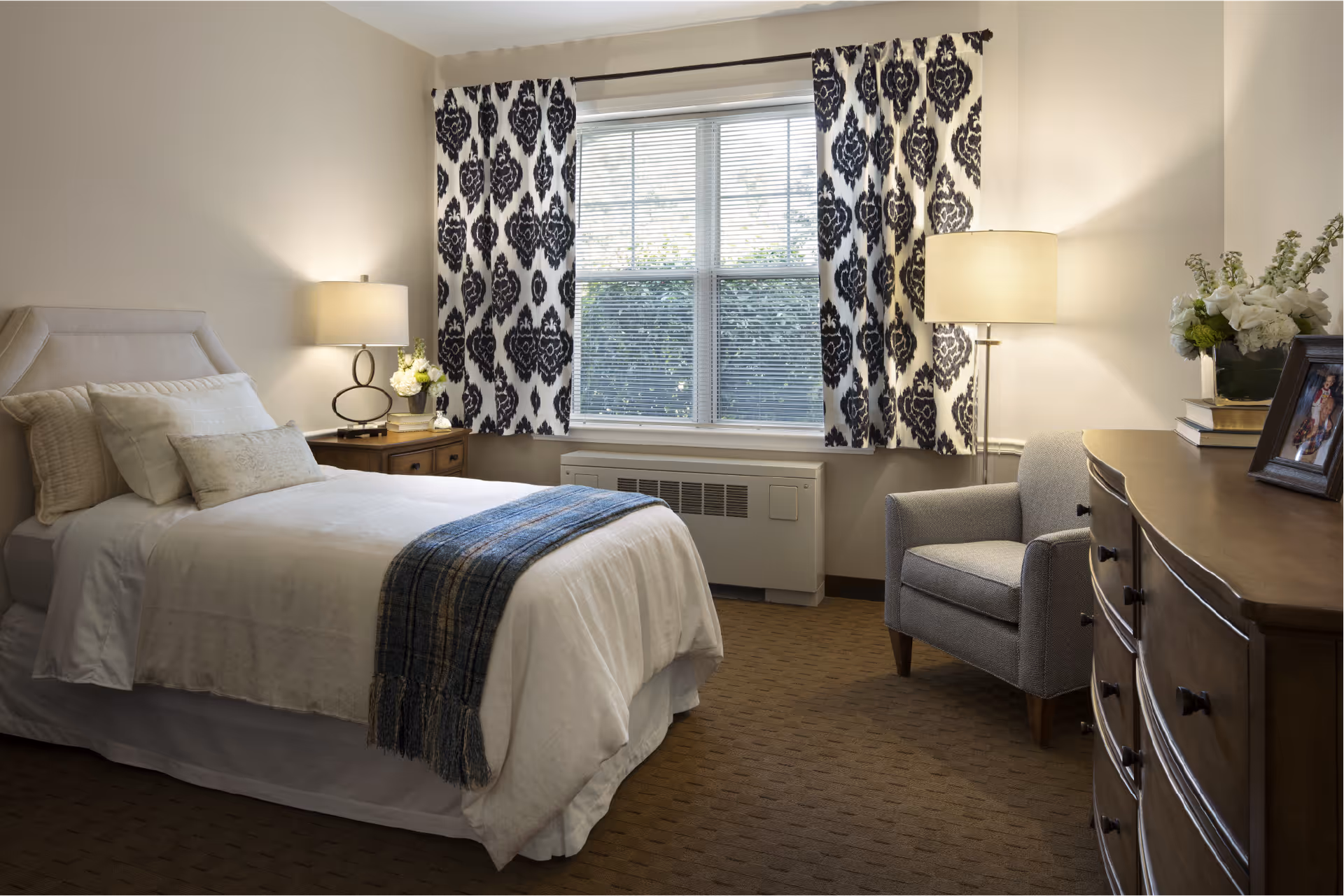 A cozy bedroom featuring a neatly made single bed with white bedding and a blue throw blanket. There is a wooden nightstand with a lamp and flowers beside the bed. A large window with patterned curtains lets in natural light. A gray armchair is placed next to a wooden dresser with a framed photo and a vase of flowers on top.
