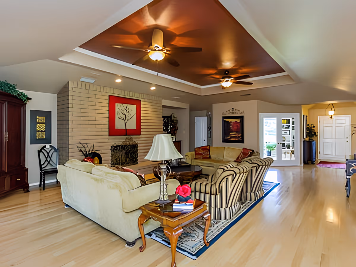 A spacious living room with light wood flooring, a beige sofa, a striped armchair, a wooden side table with a lamp, and a fireplace with a red-framed artwork above it. The ceiling has a recessed section painted in a warm brown color with two ceiling fans. There are multiple doors and windows allowing natural light into the room.