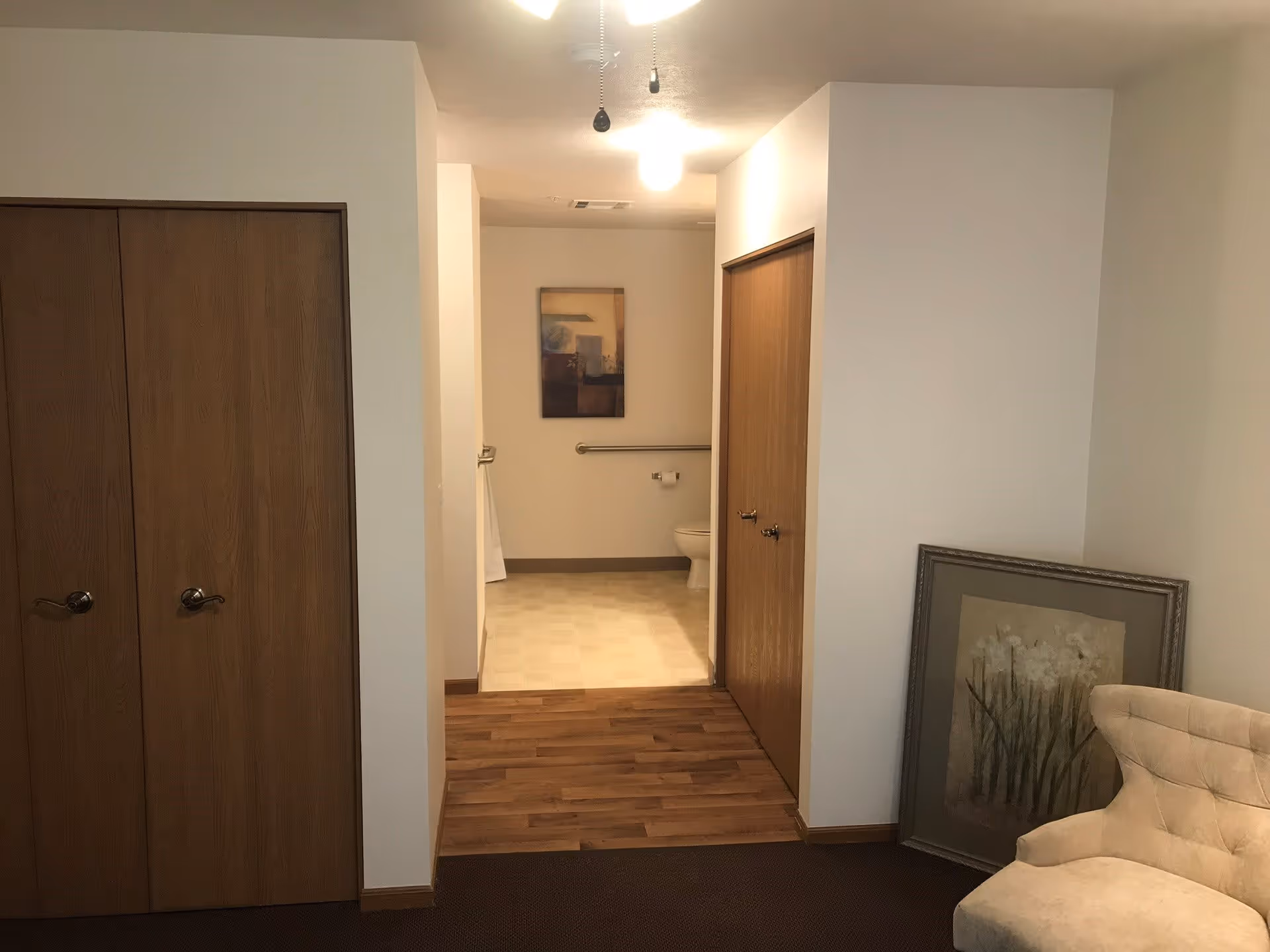 Interior view down a short hallway into a bathroom with a toilet and grab bar, flanked by wooden closet doors and a beige chair.