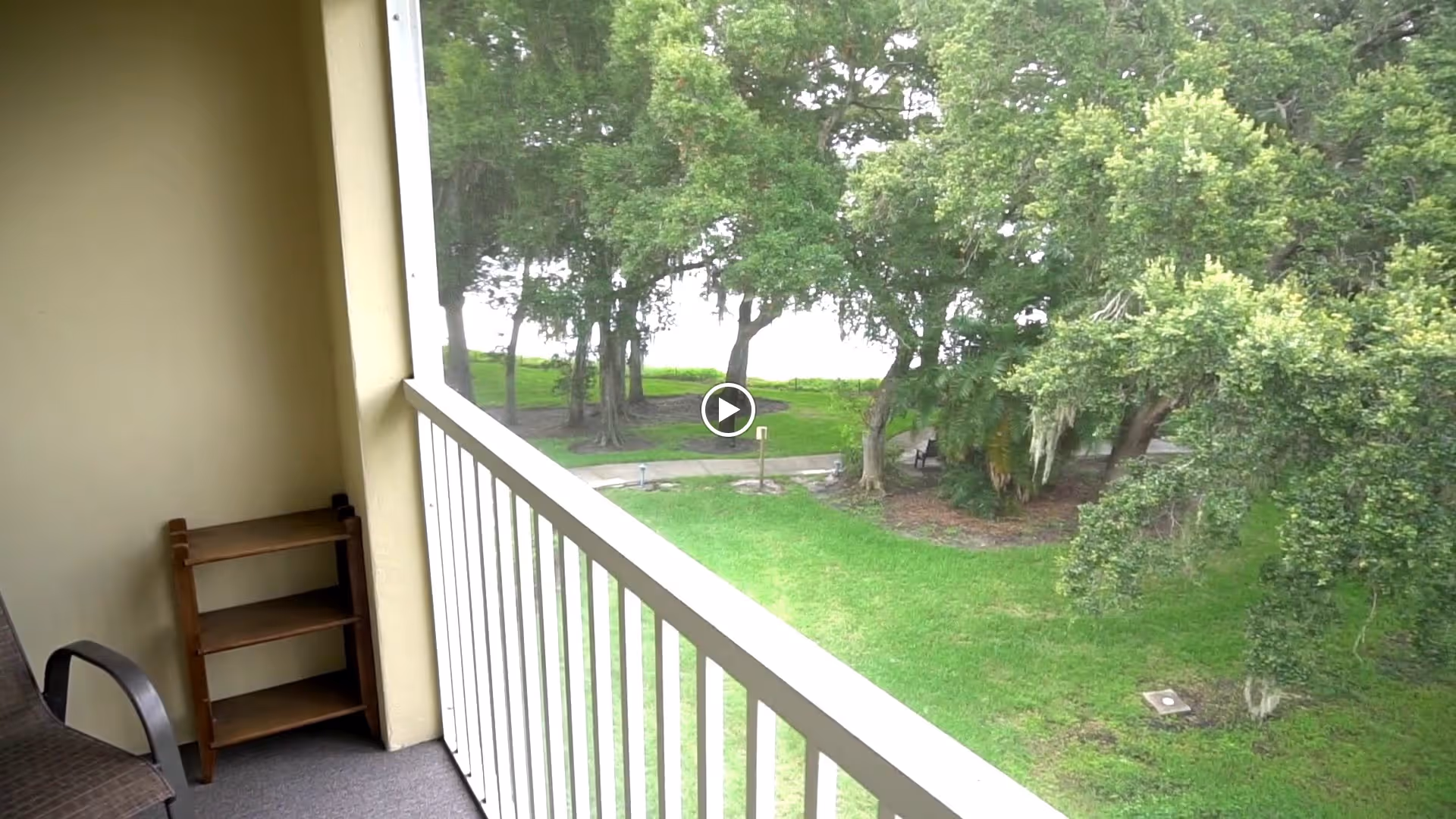 View from a balcony overlooking a green lawn with trees and a walking path. The balcony has a white railing, a brown chair, and a small wooden shelf unit against the wall.