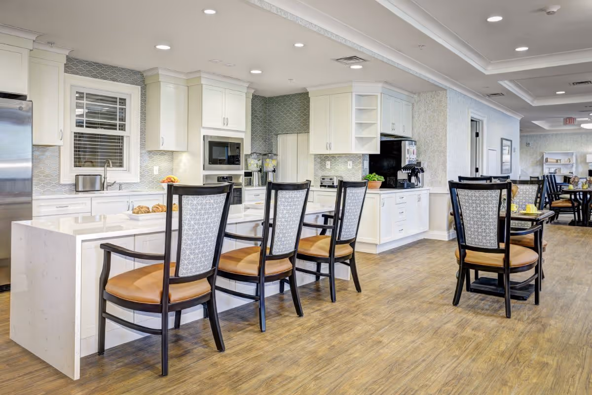 A bright and modern kitchen and dining area in a senior living facility featuring a long white island with four chairs, white cabinetry, stainless steel appliances, and a wooden floor. Additional dining tables and chairs are visible in the background.