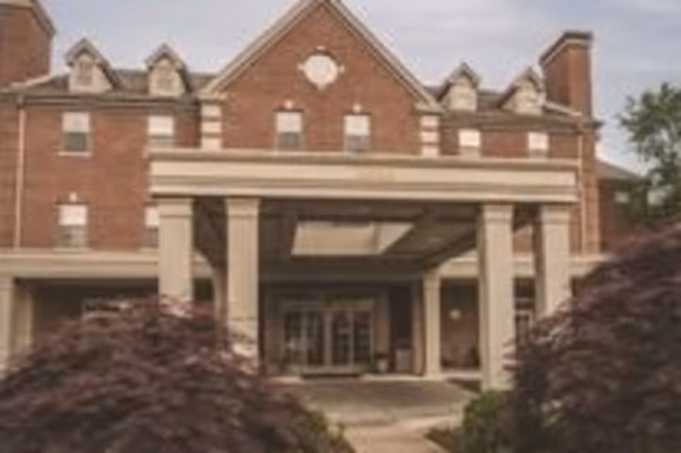 Front exterior view of a large brick building with a covered entrance supported by columns, surrounded by landscaping with bushes and trees.