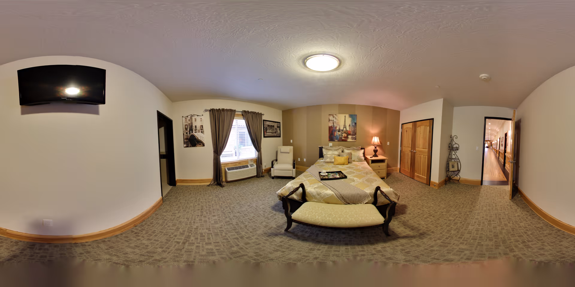 A spacious bedroom in an assisted living facility featuring a bed with a decorative bench at the foot, a nightstand with a lamp, an armchair near a window with curtains, a wall-mounted TV, and a hallway visible through an open door.