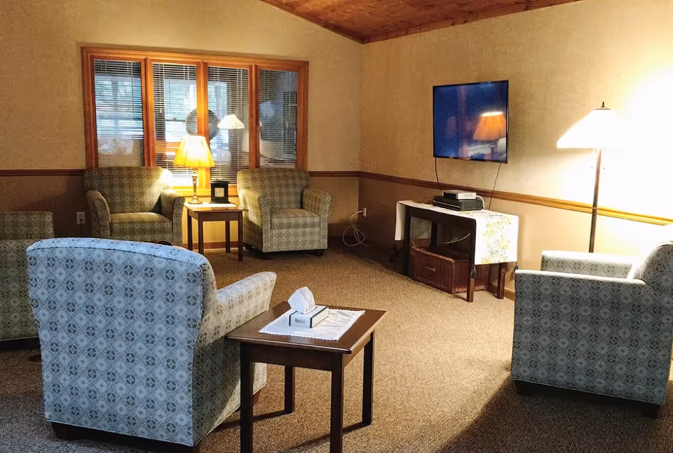 A cozy living room with patterned armchairs arranged around a small wooden table with a tissue box. There are two table lamps and a floor lamp providing warm lighting. A flat-screen TV is mounted on the wall above a table covered with a cloth, and a window with blinds is visible in the background.