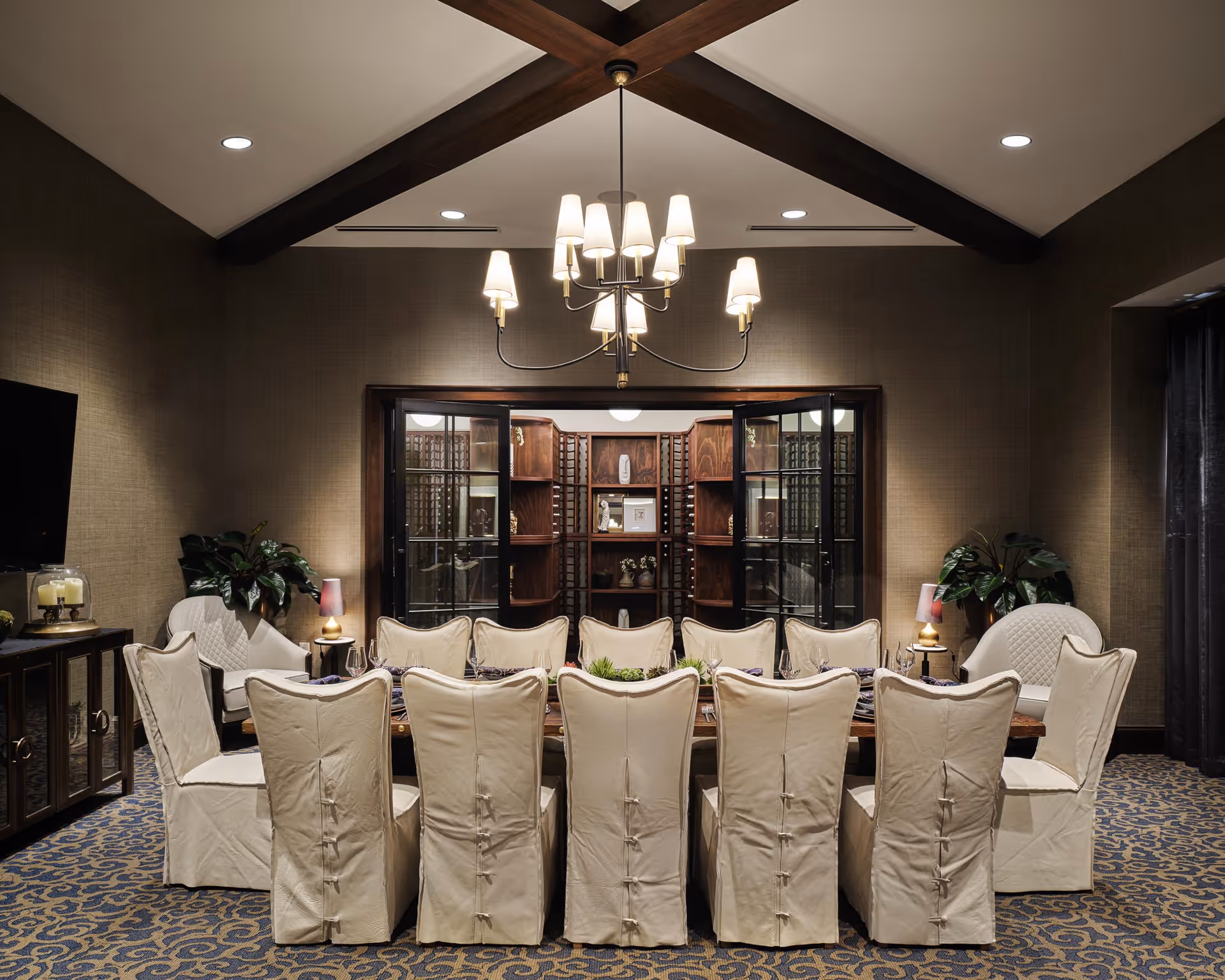 Elegant dining room with a long table surrounded by white slipcovered chairs under a chandelier, with built-in shelving and plants in the background.