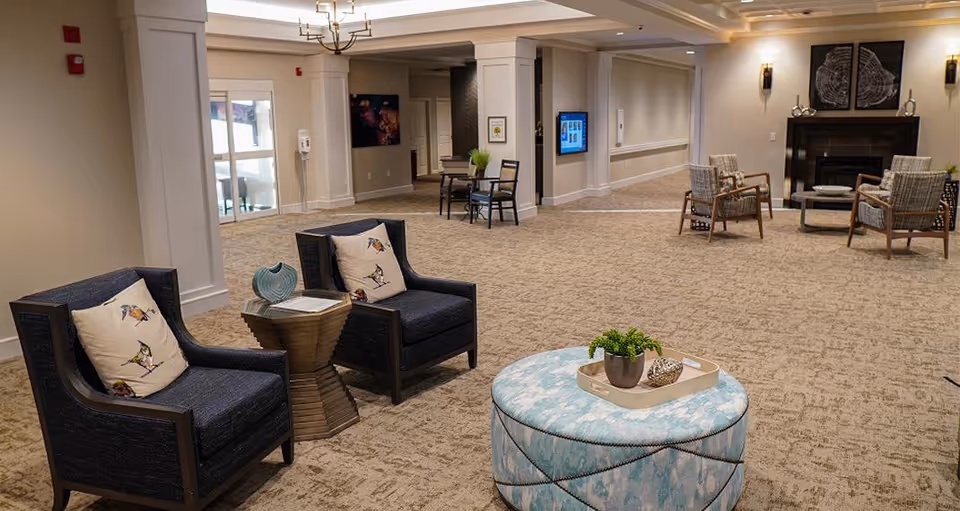 A spacious and well-lit common area in a senior living facility with beige carpet and neutral-colored walls. The room features two dark armchairs with bird-patterned cushions, a small wooden side table with decorative items, a round blue patterned ottoman with a tray holding a plant and decorative objects, and a seating area near a fireplace with two additional chairs and a coffee table. The walls have framed artwork and sconces, and there is a chandelier hanging from the ceiling.