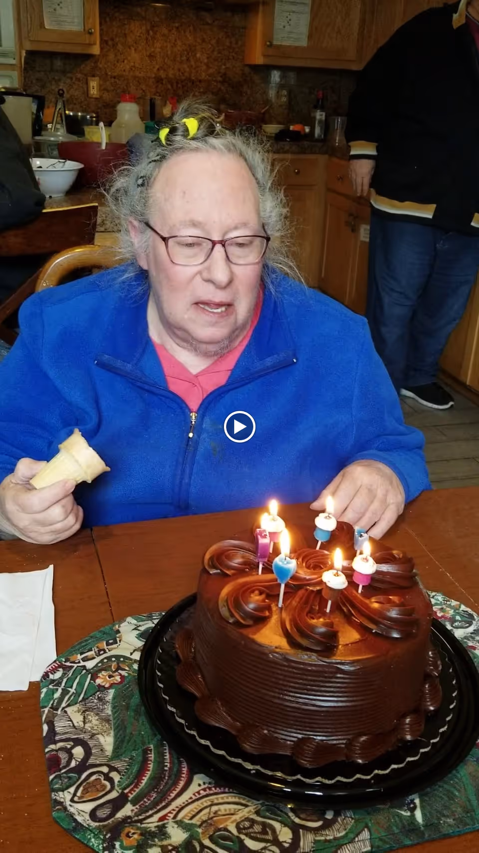 An elderly woman wearing glasses and a blue jacket is sitting at a wooden table in a kitchen. She is holding an ice cream cone in one hand and looking at a chocolate birthday cake with lit candles in front of her. The kitchen has wooden cabinets and various items on the counters. Another person is partially visible standing in the background.