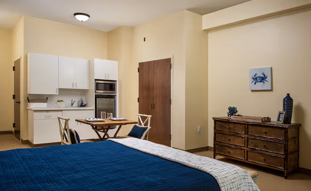 A senior living facility room featuring a bed with a blue quilt and a light-colored blanket, a small wooden dining table with two chairs, a kitchenette with white cabinets, a microwave, and a refrigerator. There is a wooden dresser with decorative items and a small painting of a blue crab on the wall.